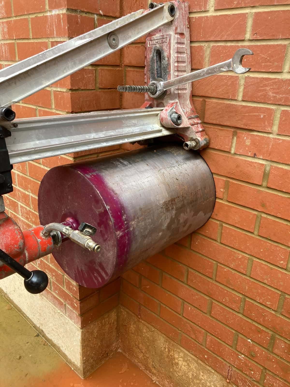 Large-diameter diamond core bit on a wall-mounted rig drilling through red brickwork at ground level