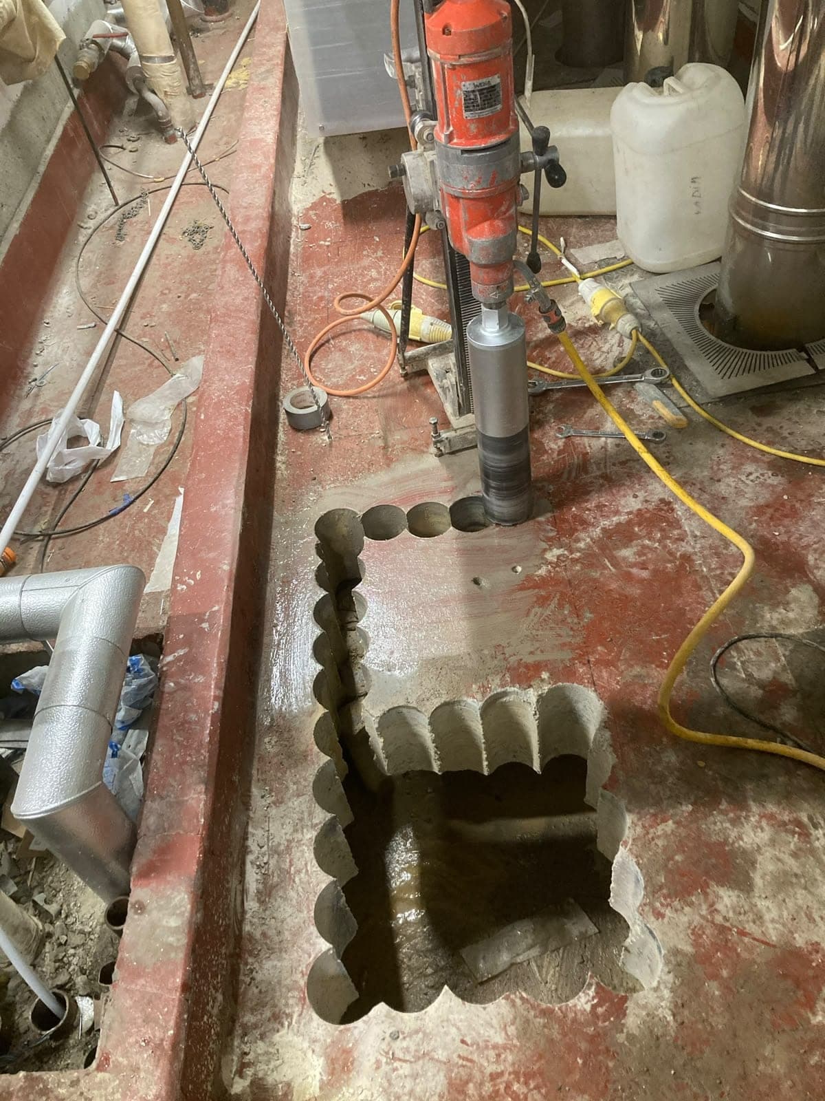 Vertical core drill rig at work on a painted concrete plant-room floor, with flue pipes alongside