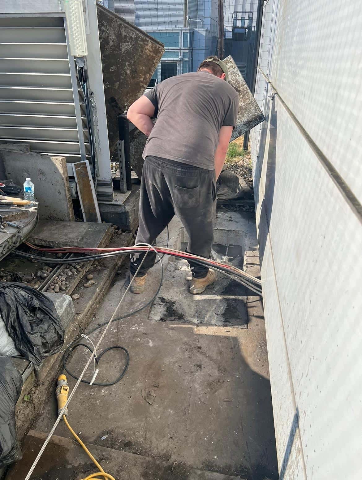 Driller working on a concrete roof walkway beside building cladding, with drill cables running along the slab