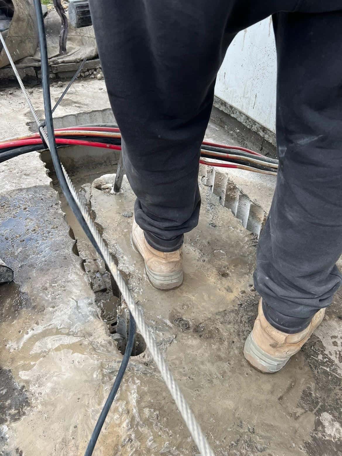 Looking down at a stitch-drilled opening in a concrete slab between the driller’s boots, with slurry on the slab