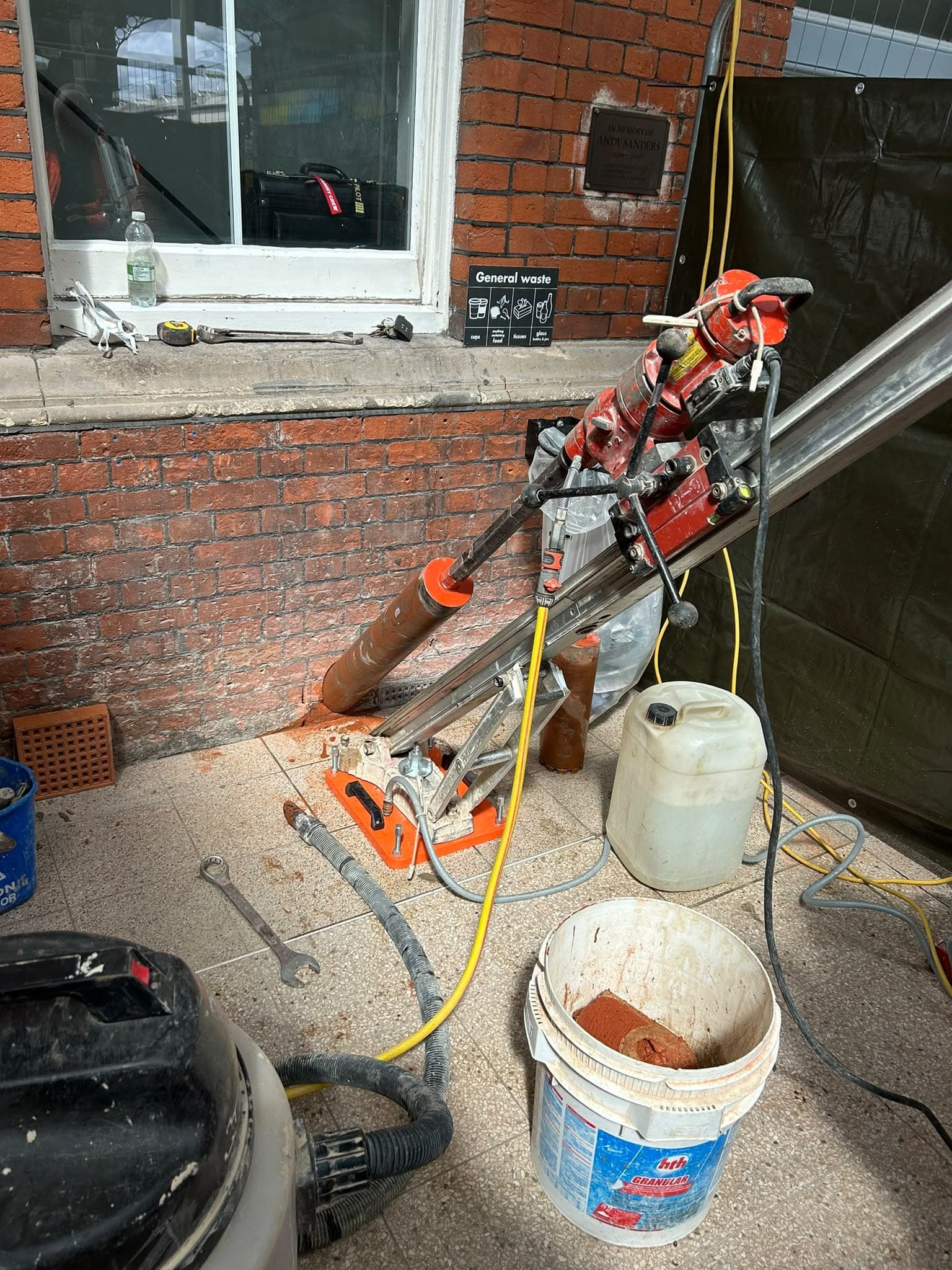 Core drill rig angled against a brick wall at floor level, with a wet vac and buckets on a tiled floor