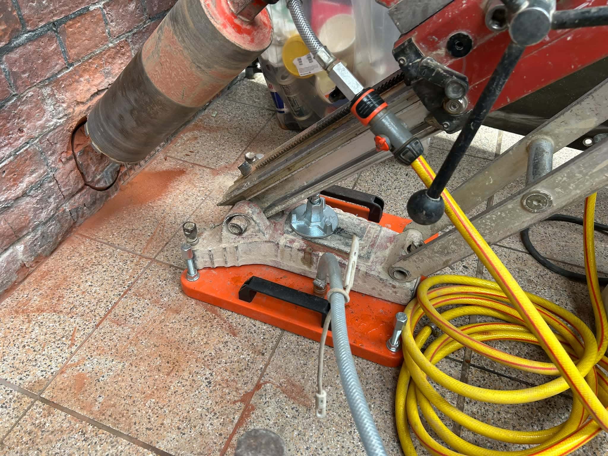 Close-up of a vacuum-base rig’s orange base plate on a tiled floor, with an angled core bit entering a brick wall