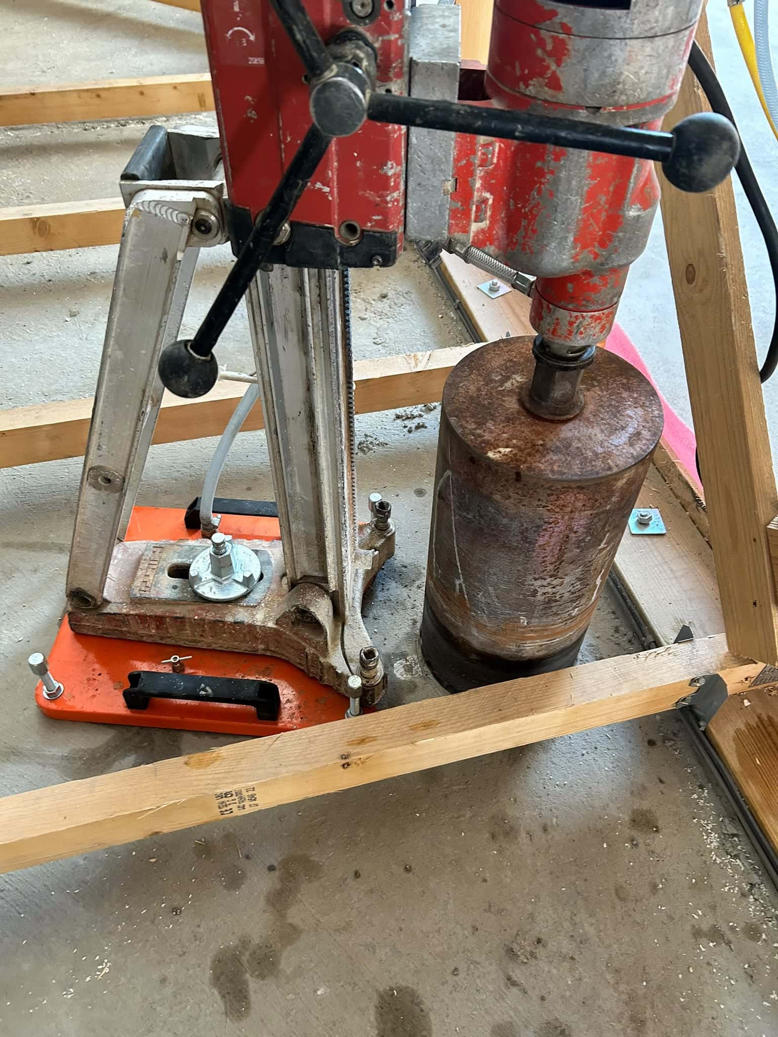 Core drill rig with a large bit drilling vertically through a floor inside a timber frame