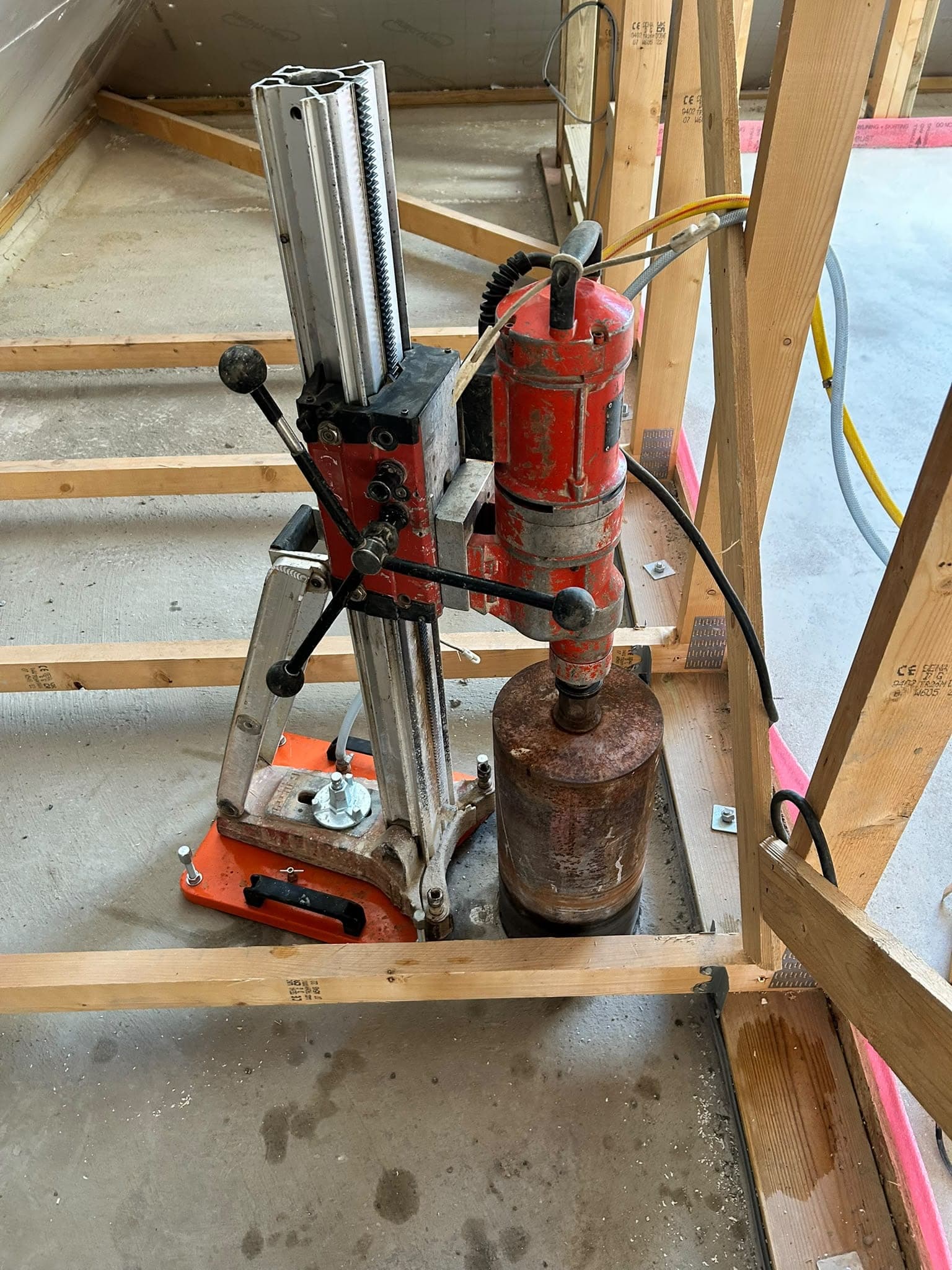 Core drill rig on an orange base plate drilling vertically through a concrete floor inside a timber frame